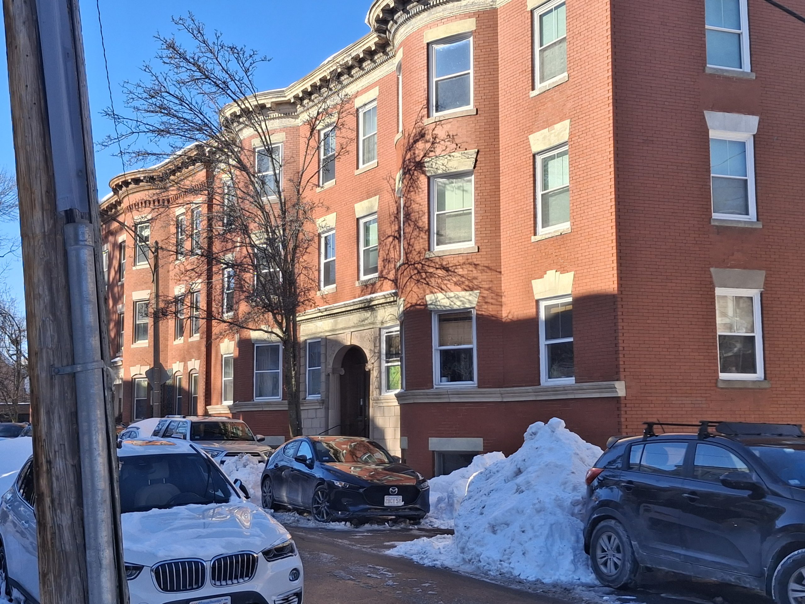 Brick condominium building on a residential street in Jamaica Plain, Boston, with parked cars and snow along the sidewalk on a clear winter day.