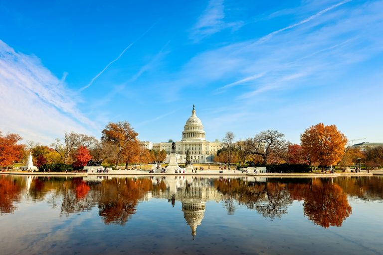 U.S. Capitol building reflected in a pool with autumn trees under a blue sky