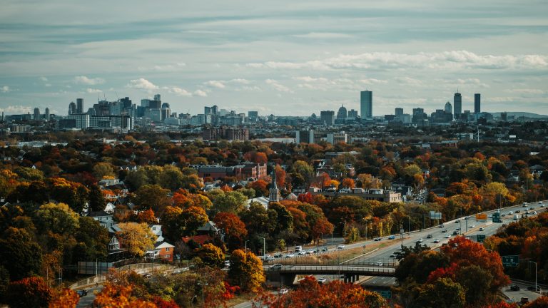 Boston skyline above Greater Boston neighborhoods, fall foliage, homes and highways illustrating the regional real estate market.