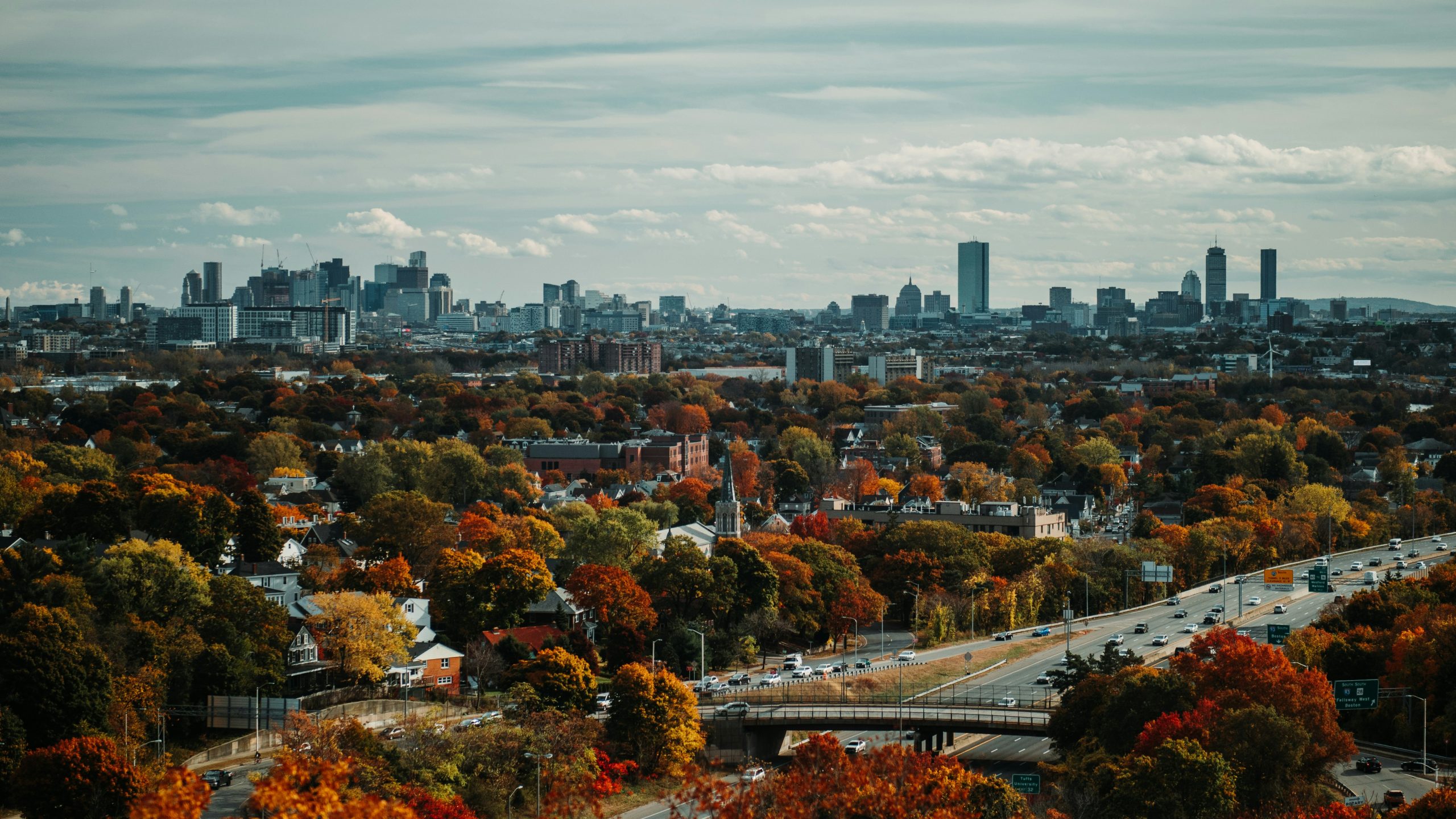 Boston skyline above Greater Boston neighborhoods, fall foliage, homes and highways illustrating the regional real estate market.
